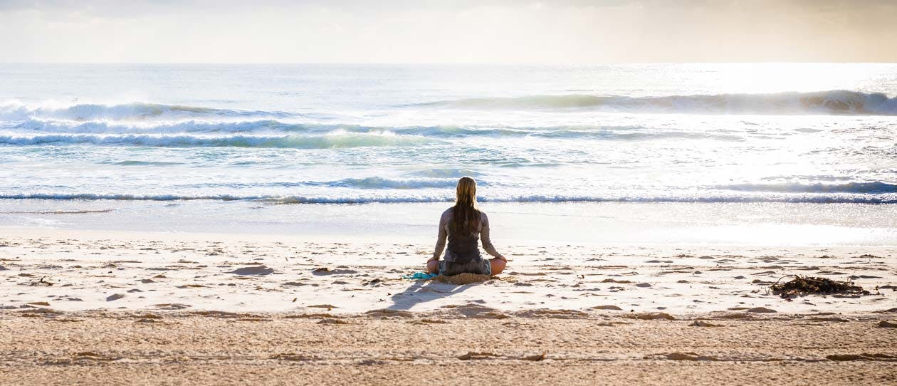 Woman sitting cross legged on the beach in the morning facing out to the ocean