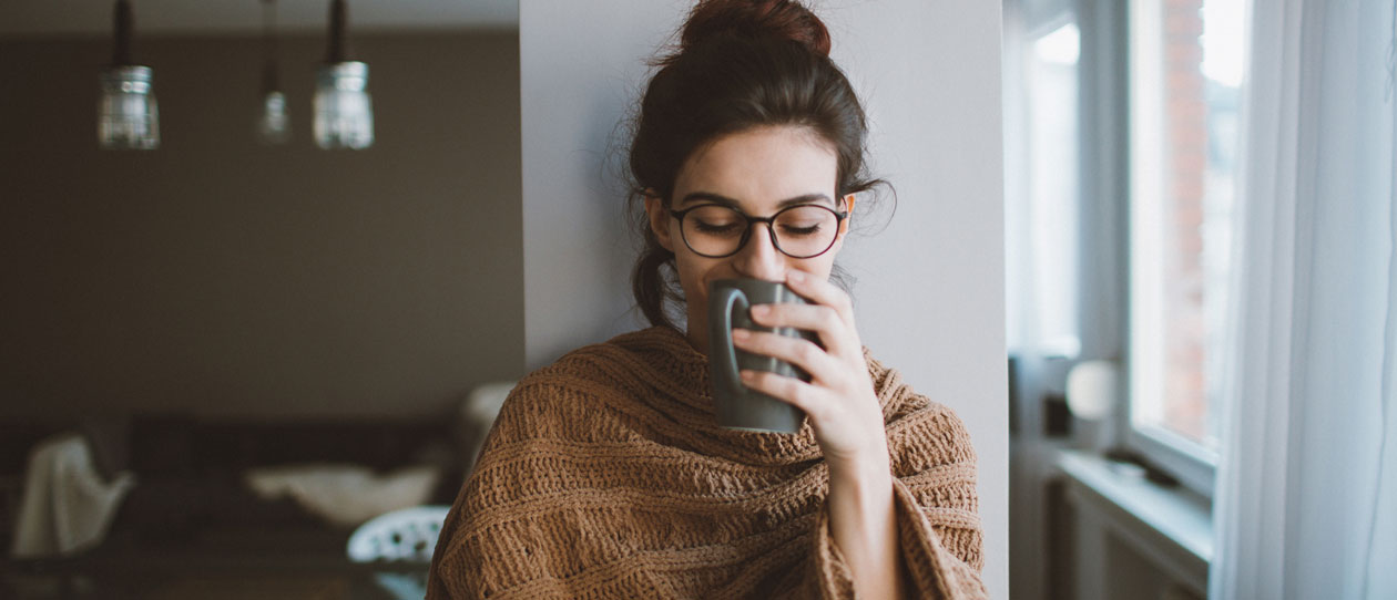  Young woman drinking her first cup of coffee early in the morning, enjoying her space and her freedom covered up in a blanket