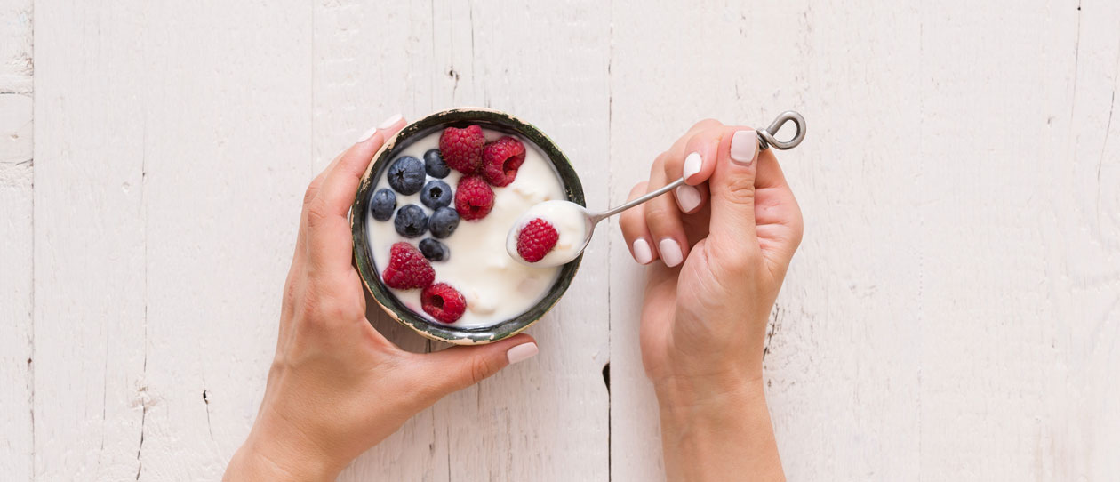 Top view on woman's hands holding small bowl with organic yogurt with blueberries and raspberries on white wooden background