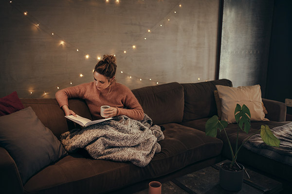 Young woman reading a book on the couch at night with a cup of tea