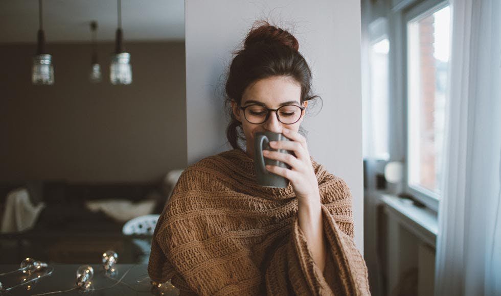  Young woman drinking her first cup of coffee early in the morning, enjoying her space and her freedom covered up in a blanket