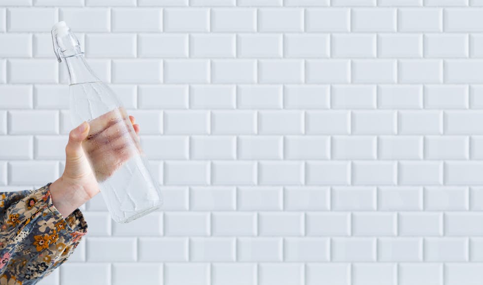 Woman holding a glass bottle of water with one hand against white subway tiles