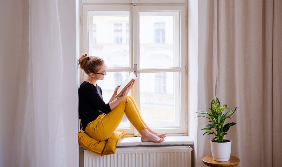 Young woman sitting in a sunlit windowsill reading a book