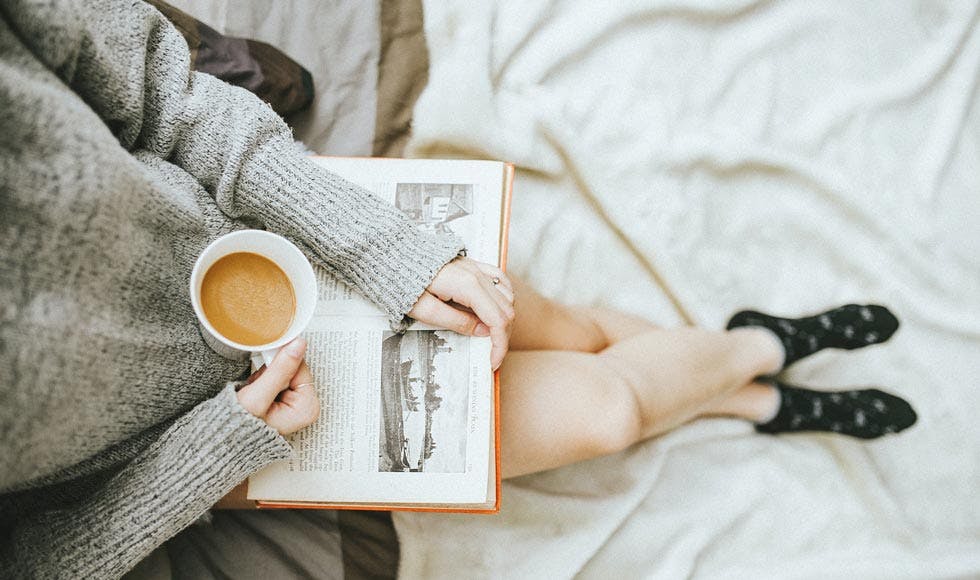 Woman holding a cup of coffee in her right hand reading a book on the bed